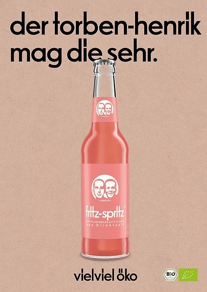 Werbeanzeige für die Bio-Schorle „fritz-spritz“ in einer Glasflasche mit rosa Etikett und dem bekannten Logo mit zwei Gesichtern. Darüber steht in schwarzer Schrift „der torben-henrik mag die sehr.“, darunter der Slogan „vielviel öko“. Im unteren rechten Bereich befinden sich zwei Bio-Siegel. Hintergrund in Kraftpapier-Optik.
