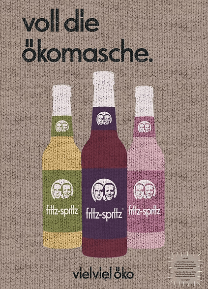Werbeanzeige für die Bio-Schorlen „fritz-spritz“ mit drei verschiedenen Glasflaschen und Etiketten mit dem jeweils bekannten Logo der zwei Gesichter der Gründer. Darüber steht in schwarzer Schrift „voll die ökomasche.“, darunter der Slogan „vielviel öko“. Im unteren rechten Bereich befinden sich zwei Bio-Siegel. Hintergrund ist ein Strick-Look.