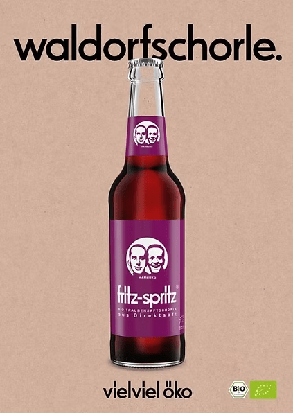 Werbeanzeige für die Bio-Traubenschorle „fritz-spritz“ in einer Glasflasche mit violettem Etikett und dem bekannten Logo mit zwei Gesichtern. Darüber steht in schwarzer Schrift „waldorfschorle.“, darunter der Slogan „vielviel öko“. Im unteren rechten Bereich befinden sich zwei Bio-Siegel. Hintergrund in Kraftpapier-Optik.
