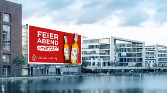 Großes rotes Werbeplakat an einem modernen Gebäude mit zwei Flaschen König Pilsener Bier und dem Text 'FEIERABEND und jetzt?'
