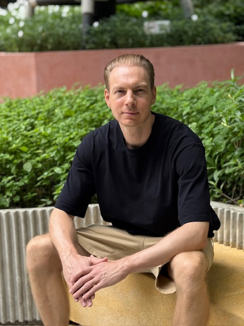 Manuel Kersting sitzt auf einer Bank. Im Hintergrund ist ein Bürokomplex in der Natur. 